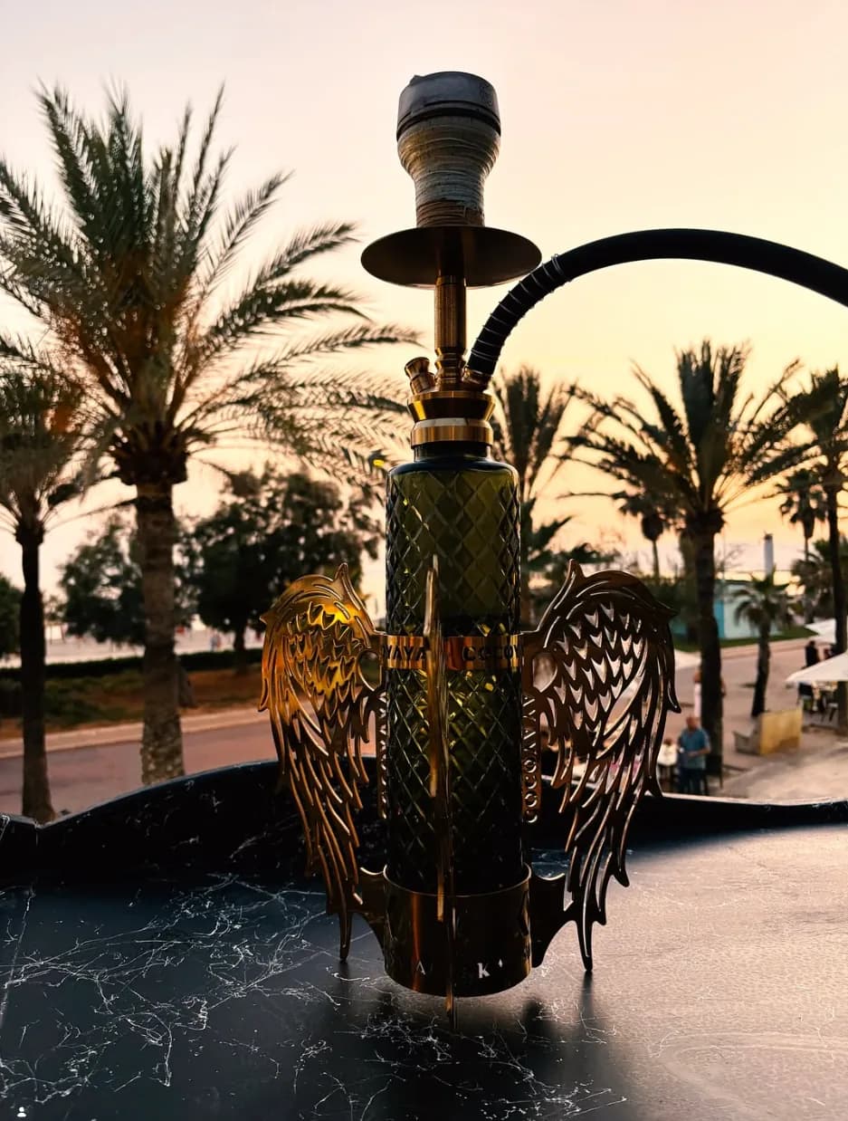 Cachimba con alas doradas sobre una mesa de mármol negro, con palmeras y el atardecer de Platja de Palma al fondo