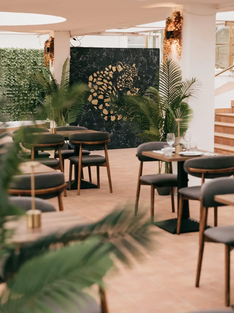 Terraza de Karisma Beach con mesas de madera, sillas de terciopelo verde y un panel de mármol negro con un felino en pan de oro
